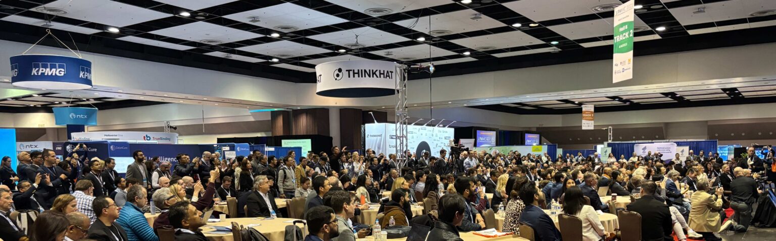 PMWC exhibition floor with attendees and booths surrounding the conference program